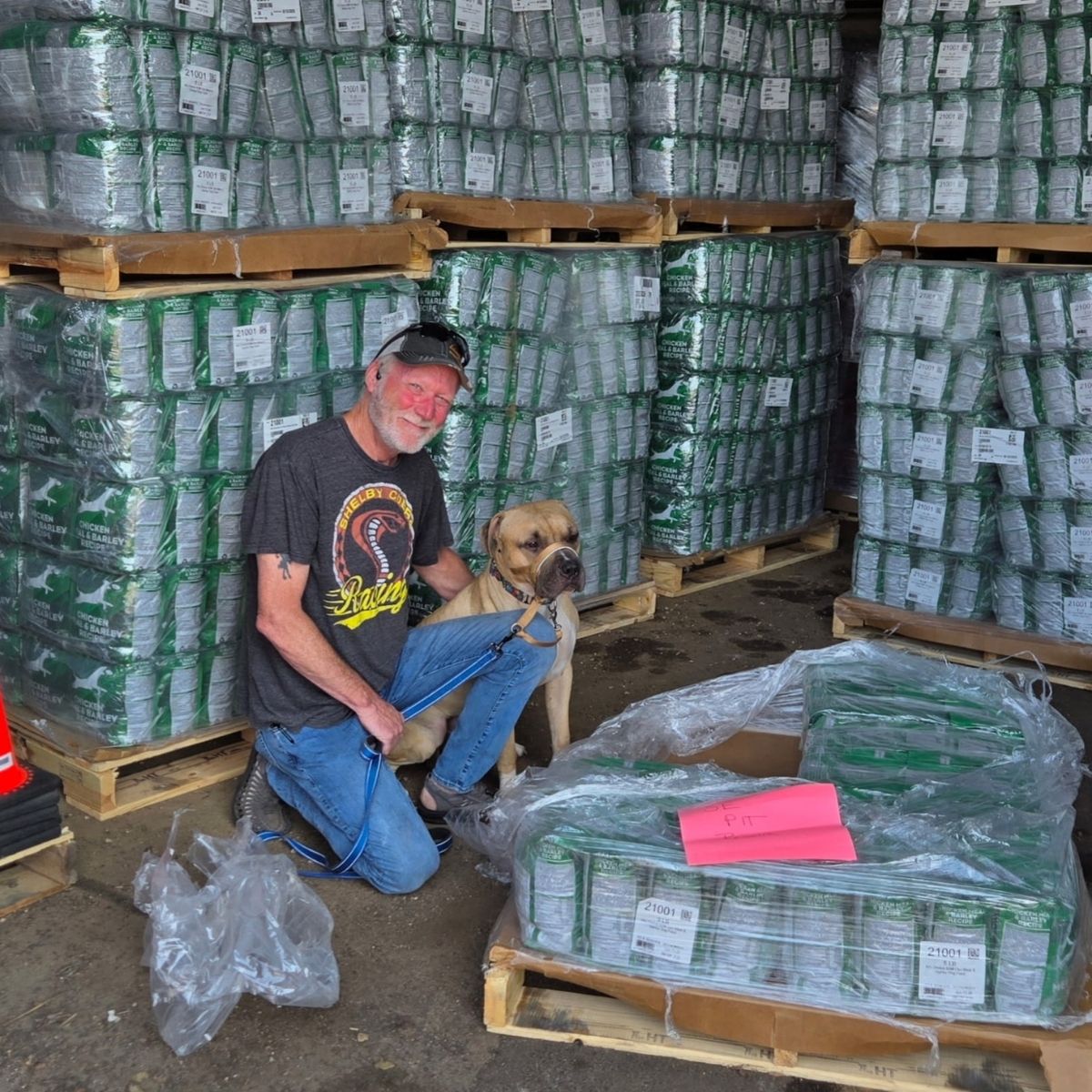 A person kneeling beside a dog among pallets of donated NutriSource dog food at Roxi & Co.
